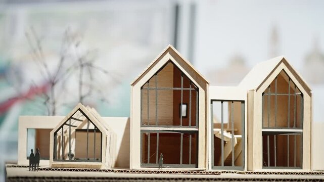 Close-up of a contemporary gabled house architectural scale model with large glass facades, wood framing, and miniature figures, on a studio table with blurred design sketches behind.