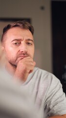 Naklejka premium Vertical photo Man pausing for reflection in mirror, gray sweater, thoughtful gaze, hand on chin, soft studio light, closeup portrait, preparation, introspective atmosphere, subtle determination