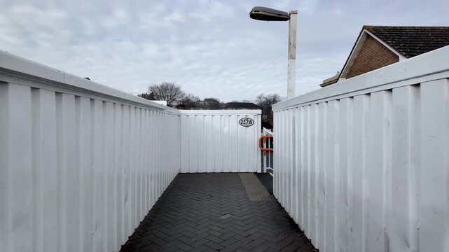 This POV shot shows a footbridge with white metal walls and a gray brick path under an overcast sky.