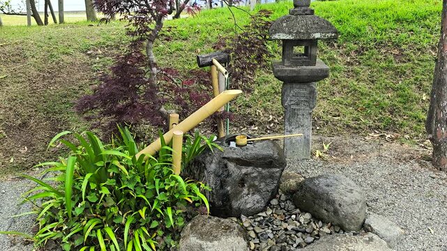 Japanese shishi odoshi bamboo fountain and tsukubai basin creating peaceful water feature in zen garden with stone lantern and lush green trees in Kumamoto, Japan