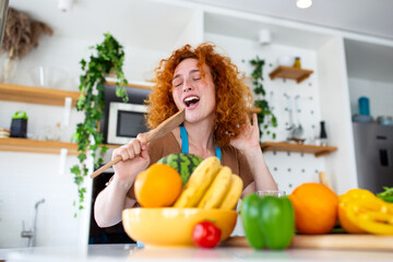 Happy redhead woman singing into a wooden spatula while cooking in the kitchen. Joyful female with...