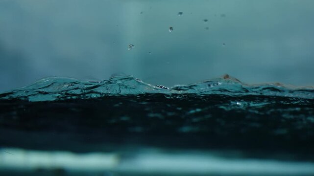 Slow-motion macro view of a dark teal water surface with delicate ripples, droplets, and gentle wave motion, capturing aquatic textures and a moody, tranquil atmosphere