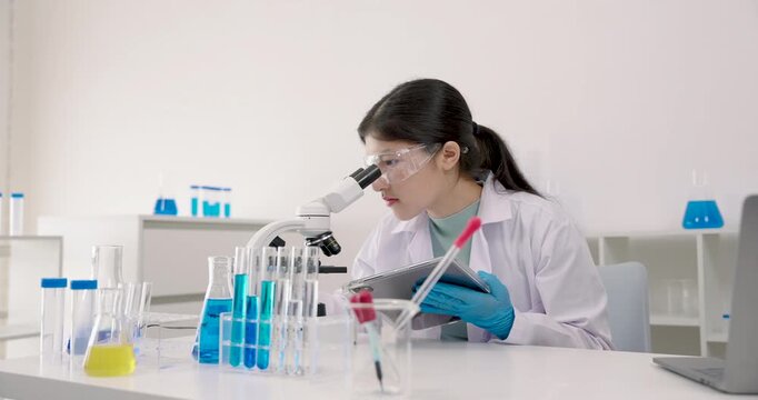Asian female student analyzing microscope slides while recording results on tablet during school laboratory experiment with various testtubes, beakers, and blue chemicals placed on white desk