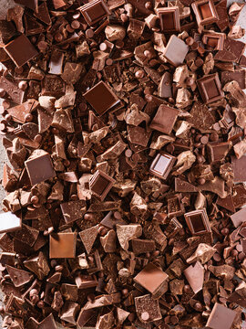Various chocolate pieces sit on a flat surface, showing chunks, chips, and squares. The different forms suggest use in baking or candy making. The color ranges from dark to light brown