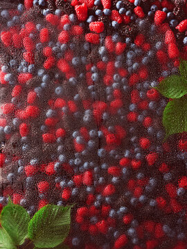 Fresh raspberries and blueberries rest on wet glass, with water droplets adding a soft look. Green mint leaves enhance the scene. This captures summer freshness and healthy ingredients