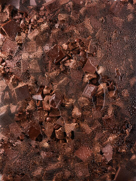 Assorted chocolate pieces rest behind wet glass with fine droplets. The scene includes broken bars and chocolate pearls in deep brown tones, showcasing richness and depth
