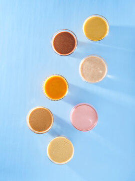 Eight glasses filled with various colored drinks are set on a light blue surface. The drinks range from orange to pink and brown. Each glass shows a different texture and consistency