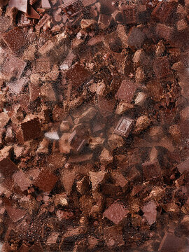 Assorted chocolate bars, shards, and pearls are placed behind a wet glass. Water droplets distort the view, creating depth and showcasing the rich texture of the chocolate