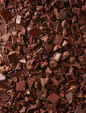Small chocolate pieces and chips are spread out on a flat surface. They vary in shape and size and are ready for use in recipes. This collection shows bits of dark chocolate