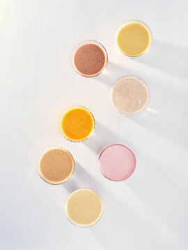 Several clear cups hold different types of smoothies. These cups are placed on a white surface and are arranged in a circle. The light reflects on the cups and the surface