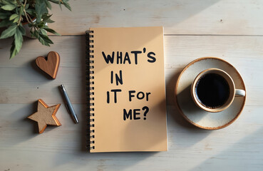 Fototapeta premium Notebook with question Whats in it for me next to coffee cup. Pen and wooden heart star decor on desk. Morning light streams across wooden table. Inspiration for goal setting.