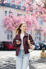 Young woman walking in spring city with smartphone. Girl enjoying cherry blossoms on sunny street with blooming trees, pink flowers. Active urban lifestyle with movement.