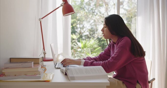 Young Asian teen types on a silver laptop at a tidy home desk with books, coral lamp, and sheer curtains
