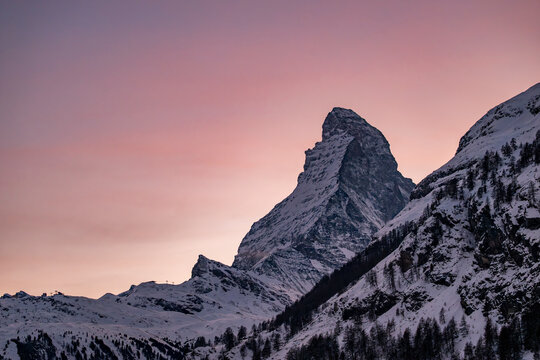 Matterhorn peak at sunset with a pink sky, snow covered alpine mountains and winter terrain near Zermatt ski resort in Switzerland.