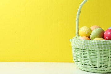 Painted Easter eggs in wicker basket on white table against yellow background, closeup. Space for text