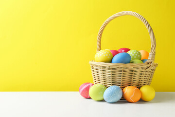 Painted Easter eggs in wicker basket on white table against yellow background. Space for text