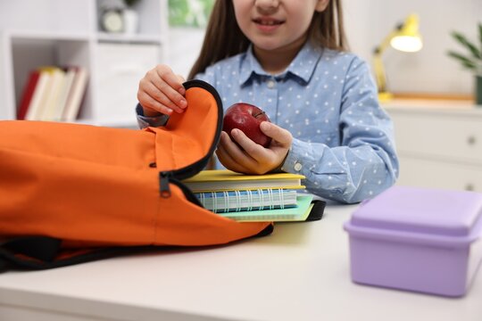 Girl with apple packing her backpack for school at desk indoors, closeup