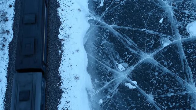 A dark train moves continuously along tracks bordering a vast, intensely cracked frozen body of water, seen from an aerial perspective in a cold, stark environment.