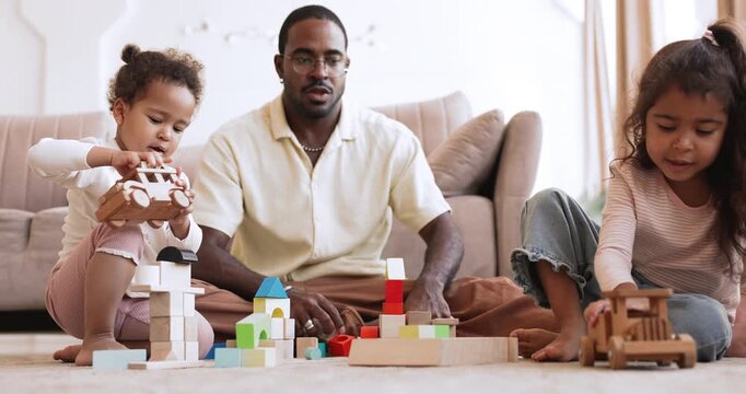 Loving black father sitting on floor and playing wooden toys and blocks with two little biracial daughters. Developmental games with kids at home, fatherhood, family playtime in modern living room