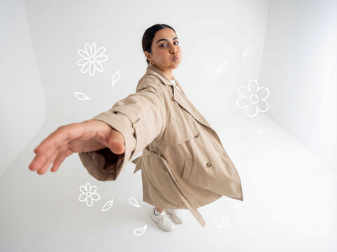 Young middle eastern woman in oversized trench coat posing wacky fisheye lens perspective high angle white studio background white doodle elements fashion advertisement copy space