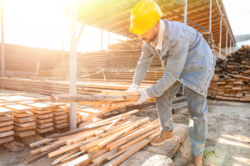 Fototapeta premium Male carpenter in hardhat with wooden boards working at sawmill