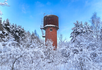 Old red brick water tower in snowy winter forest.Vintage red brick water tower surrounded by snow-covered trees under a bright blue winter sky