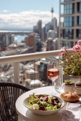 Naklejka premium Light lunch on sunlit balcony table: salad and glass of rose, distant city skyline softened by spring haze. A moment of quiet refreshment with urban charm.
