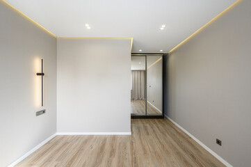 Obraz premium Modern minimalist room with light wooden flooring, neutral walls, recessed ceiling lights, LED strip, sleek wall lamp, mirrored wardrobe, clean lines, spacious layout.