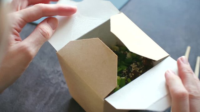 Close up female hands opening cardboard box with delicious takeout meal inside, revealing fresh dish of wok noodles, rice garnished with spring onions, sesame seeds. Takeout container with asian food