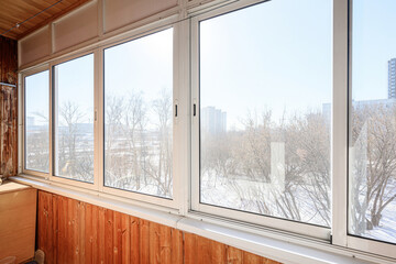Obraz premium Balcony with large white-framed windows, wooden paneling, view of snowy landscape, bare trees, distant buildings; bright, airy, cozy winter setting.