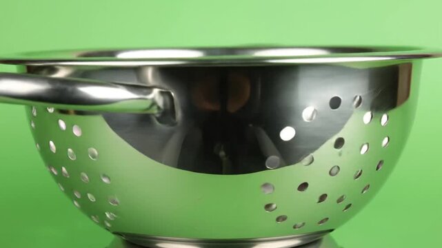 Close-up of a stainless steel kitchen colander on a green background. Handles and perforations are visible