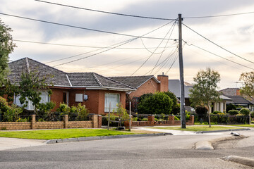 A quiet suburban residential street with single-storey brick houses, front gardens, and overhead power lines at a street corner. Calm neighborhood, housing, local infrastructure, peaceful living © Doublelee