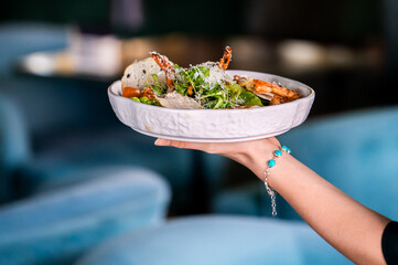 A hand holding a white bowl filled with a gourmet shrimp and green salad with croutons, parmesan, and crab claws. It is shot in a restaurant setting with a shallow depth of field and blue background. © pavel siamionov