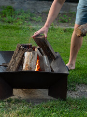Man Adjusting Firewood in Metal Fire Pit with Flames in Green Backyard
