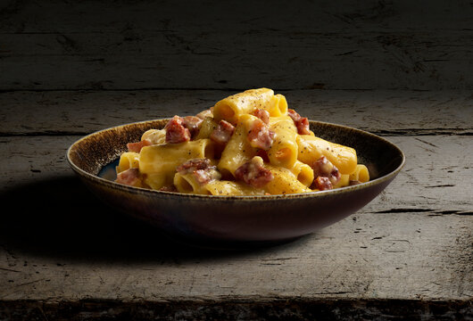 Dark bowl with rigatoni pasta carbonara on rough wood