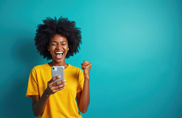 Young african american woman cheers happily holding smartphone. She expresses joy and excitement, raising fist in triumph. Winning moment captured using mobile tech.