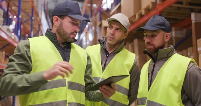 Workers supervisors discuss logistics in bright warehouse. Wearing safety vests, the team reviews planning on a tablet, with a supervisor pointing at racks. Teamwork keeps logistics on track.