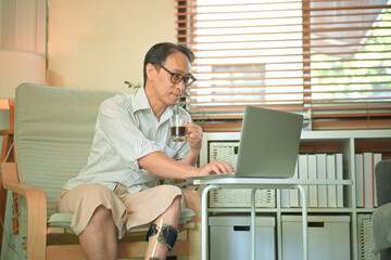 Middle-aged man working on a laptop at home while wearing a prosthetic leg, focused remote work