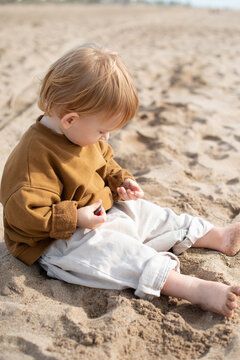 Toddler sitting on sandy beach and playing with sand in hands. Soft natural light, warm tones, and candid moment capture curiosity, childhood innocence, and outdoor exploration. Ideal for concepts of 
