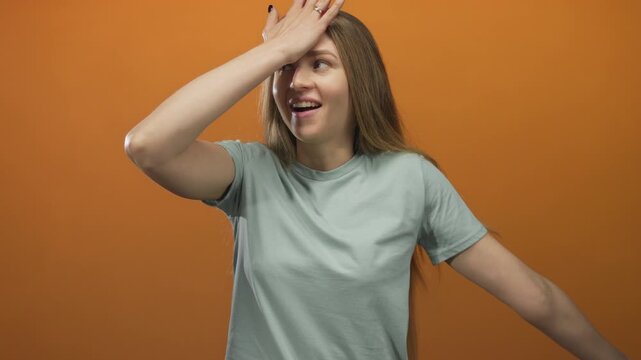 Woman facepalming in a blue shirt against an orange background, expressing frustration and surprise with bright isolated setting highlighting emotion and gesture.