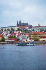 Obraz premium A scenic Prague riverside captures colorful rooftops, a modern boat gliding on the river, and iconic cathedral spires overlooking the historic cityscape of the Czech Republic.