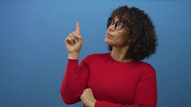 Hispanic woman wearing glasses and red sweater pointing finger with folded arm in studio against a blue colored background; confidence.