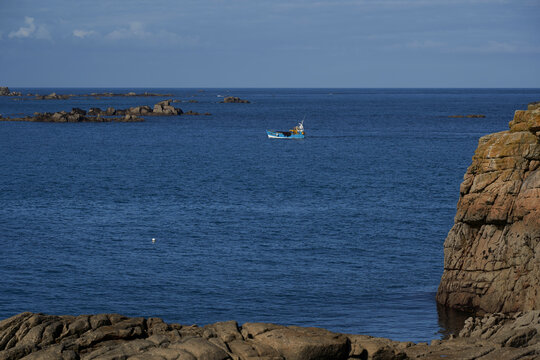 A fishing boat sails on the blue sea, surrounded by rocky islands and cliffs, showcasing the serene beauty of Brittany. Plage des Amiets, France.
