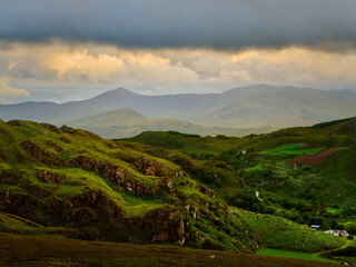 Obraz premium Verdant hillside descends into a valley in National Park with distant mountains and moody, overcast sky.
