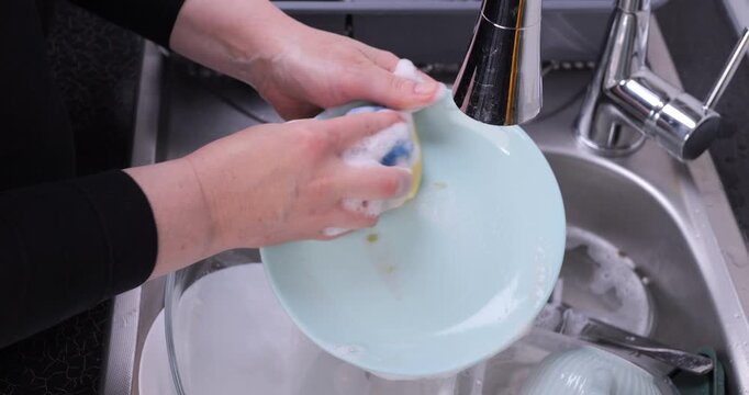 Close-up of a hand washing a plate with sponge, soap, and water in a sink full of dishes. Dish washing, kitchen cleaning, and household hygiene concept.