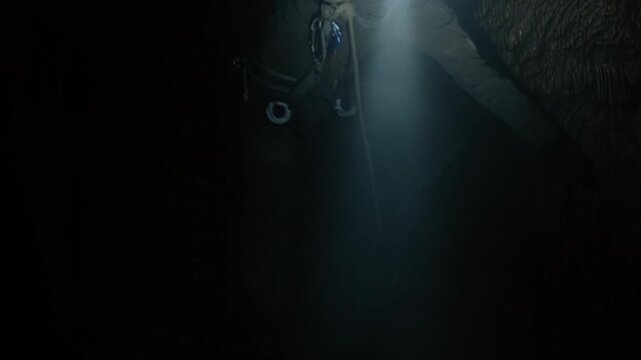 Man rappelling down into dark limestone cave. Bright headlamp light, cinematic misty atmosphere. Speleology concept.