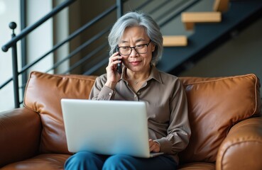 Elderly Asian woman wearing glasses talks on cell phone while using laptop. She sits on brown leather couch in modern living room near stairs. She appears focused on work.