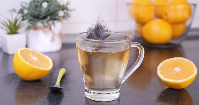 Close-up of a tea bag being dipped and lifted in hot water inside a mug on a kitchen countertop, with lemons nearby. Tea infusion process, cozy and calming moment.