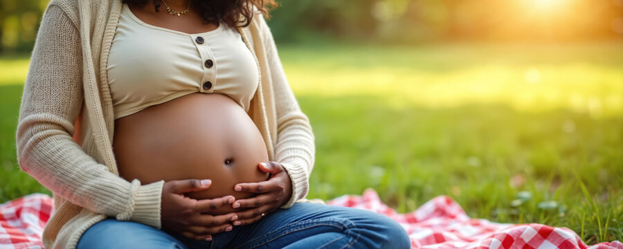 Young african american woman pregnant belly sitting outdoors on checkered blanket. Gentle hands caress tummy, warm sunshine. Maternity lifestyle outdoor relaxation, expecting mother happy.
