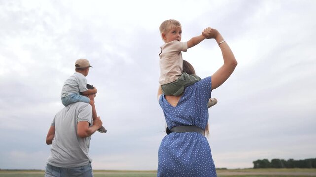 A woman with a child on her shoulders walks in a field, while a man with a child on his shoulders walks alongside. The woman holds the child's hands, and the man holds the other ch